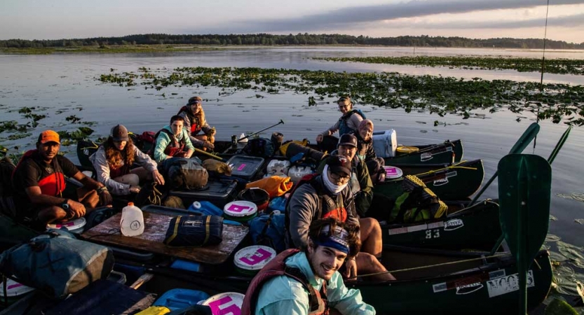 A group of people sitting in canoes that are tied together smile at the camera. They appear to be floating in shallow water. 